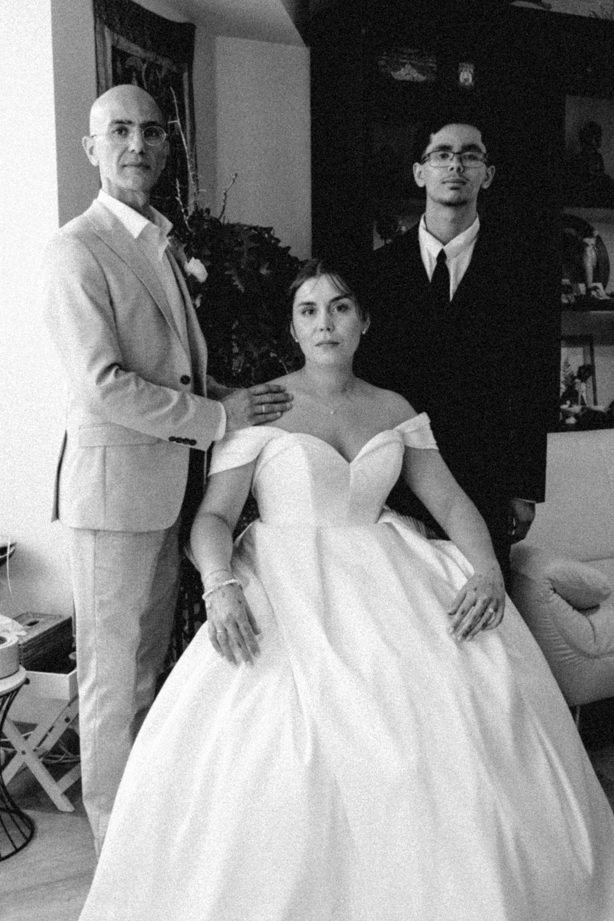 Bride seated, with the groom and a young man standing behind her, in black and white